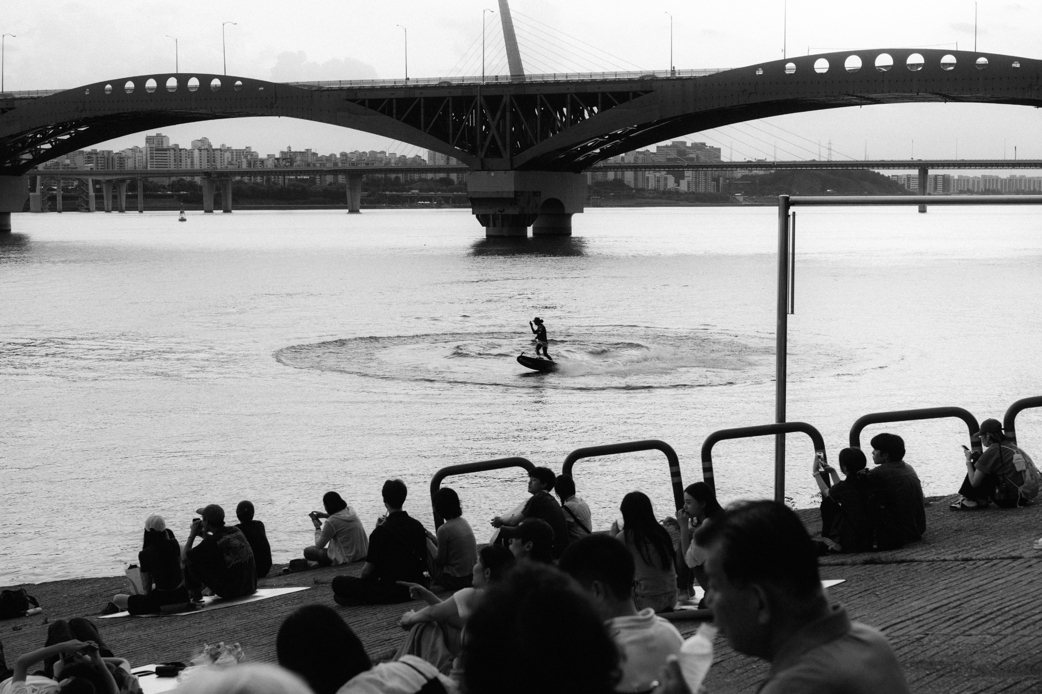 People gathered beside a river beneath an arched bridge in black and white