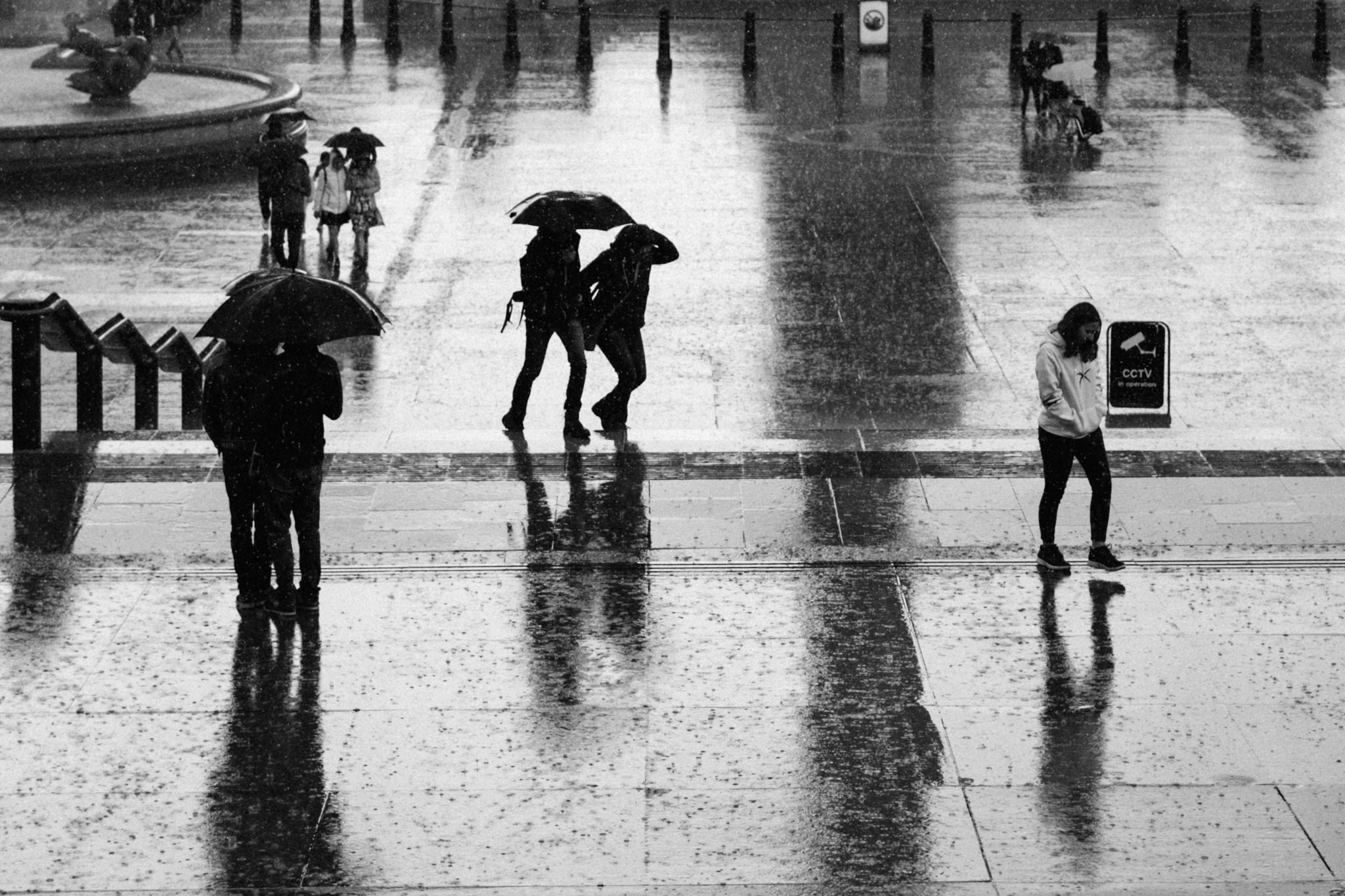 Pedestrians with umbrellas crossing a rain-soaked square in black and white